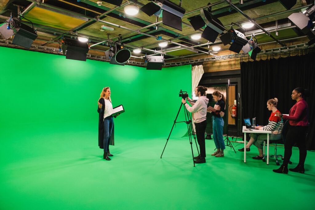 A wide-angle shot of a multi-ethnic group of people working in a film studio, a mature caucasian woman can be seen presenting in front of a green screen.