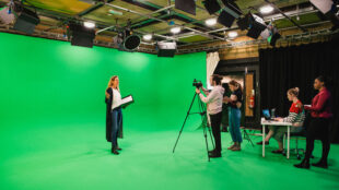 A wide-angle shot of a multi-ethnic group of people working in a film studio, a mature caucasian woman can be seen presenting in front of a green screen.