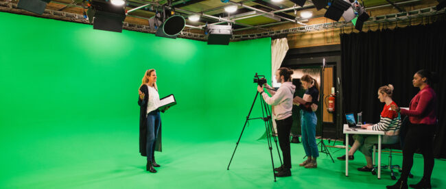 A wide-angle shot of a multi-ethnic group of people working in a film studio, a mature caucasian woman can be seen presenting in front of a green screen.