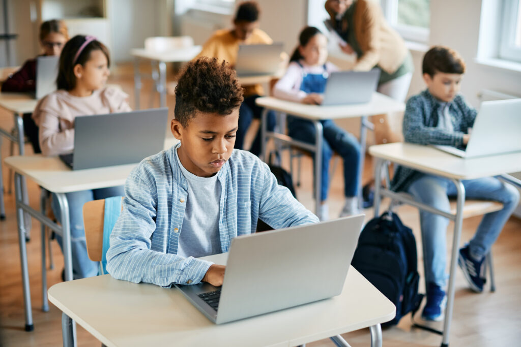 A classroom with students sitting at desks, using laptops. In the foreground, a boy is focused on his laptop, with other students in the background also working on their computers. The environment appears to be a modern, technology-enabled learning space.