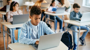 A classroom with students sitting at desks, using laptops. In the foreground, a boy is focused on his laptop, with other students in the background also working on their computers. The environment appears to be a modern, technology-enabled learning space.