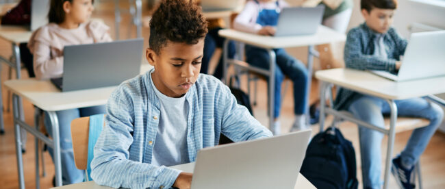 A classroom with students sitting at desks, using laptops. In the foreground, a boy is focused on his laptop, with other students in the background also working on their computers. The environment appears to be a modern, technology-enabled learning space.