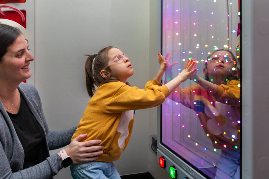 A young student in a yellow sweatshirt, wearing glasses and what appears to be a hearing aid, reaches out to interact with a colorful light display panel while being supported by an adult in a grey sweater. The panel shows multiple colored LED lights creating an engaging sensory experience, and the student's reflection can be seen in the panel. The student appears engaged and fascinated by the interactive light display.