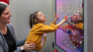 A young student in a yellow sweatshirt, wearing glasses and what appears to be a hearing aid, reaches out to interact with a colorful light display panel while being supported by an adult in a grey sweater. The panel shows multiple colored LED lights creating an engaging sensory experience, and the student's reflection can be seen in the panel. The student appears engaged and fascinated by the interactive light display.