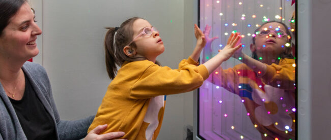 A young student in a yellow sweatshirt, wearing glasses and what appears to be a hearing aid, reaches out to interact with a colorful light display panel while being supported by an adult in a grey sweater. The panel shows multiple colored LED lights creating an engaging sensory experience, and the student's reflection can be seen in the panel. The student appears engaged and fascinated by the interactive light display.