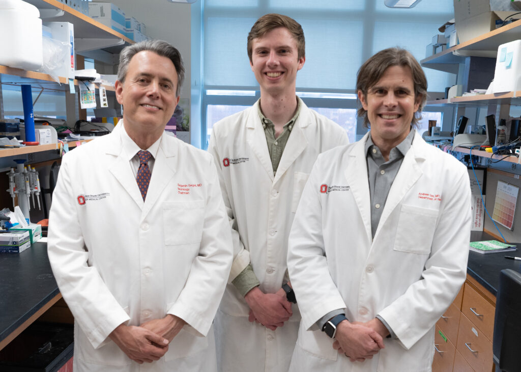 Benjamin Segal, MD, Andrew Jerome, PhD and Andrew Sas, MD, PhD, wearing white lab coats standing in what appears to be a research laboratory. They are positioned in front of laboratory shelving containing various supplies and equipment. All three are smiling at the camera, and there are Ohio State University logos visible on their lab coats.