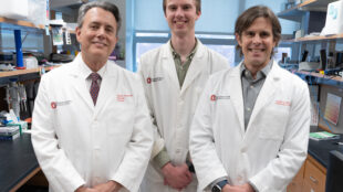 Benjamin Segal, MD, Andrew Jerome, PhD and Andrew Sas, MD, PhD, wearing white lab coats standing in what appears to be a research laboratory. They are positioned in front of laboratory shelving containing various supplies and equipment. All three are smiling at the camera, and there are Ohio State University logos visible on their lab coats.