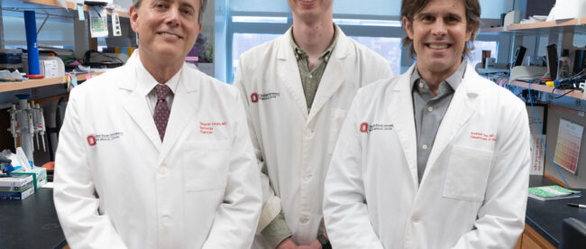 Benjamin Segal, MD, Andrew Jerome, PhD and Andrew Sas, MD, PhD, wearing white lab coats standing in what appears to be a research laboratory. They are positioned in front of laboratory shelving containing various supplies and equipment. All three are smiling at the camera, and there are Ohio State University logos visible on their lab coats.