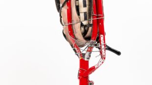 A studio photograph of the "Bike Leg" prosthetic on a white background. The leg features a red bicycle frame for its main structure, with a woven socket at the top made of tan straps and black tire pieces. The foot is a curved section of a bicycle wheel with black tire tread, and it is connected to the ankle area by a network of bicycle spokes.