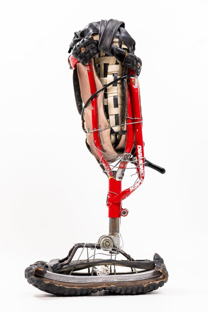 A studio photograph of the "Bike Leg" prosthetic on a white background. The leg features a red bicycle frame for its main structure, with a woven socket at the top made of tan straps and black tire pieces. The foot is a curved section of a bicycle wheel with black tire tread, and it is connected to the ankle area by a network of bicycle spokes.