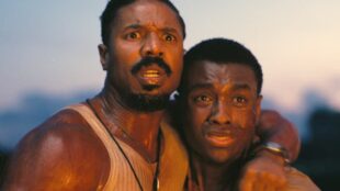 A close-up shot from the film ‘Sinners,’ featuring Michael B. Jordan and Miles Caton. Jordan, on the left with a beard and wearing a white tank top, holds Caton close. Both men have expressions of intense shock and fear on their faces as they stare at something off-camera. They are illuminated by a warm, orange glow against a dim background, suggesting they are witnessing a fire or explosion.