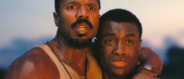 A close-up shot from the film ‘Sinners,’ featuring Michael B. Jordan and Miles Caton. Jordan, on the left with a beard and wearing a white tank top, holds Caton close. Both men have expressions of intense shock and fear on their faces as they stare at something off-camera. They are illuminated by a warm, orange glow against a dim background, suggesting they are witnessing a fire or explosion.