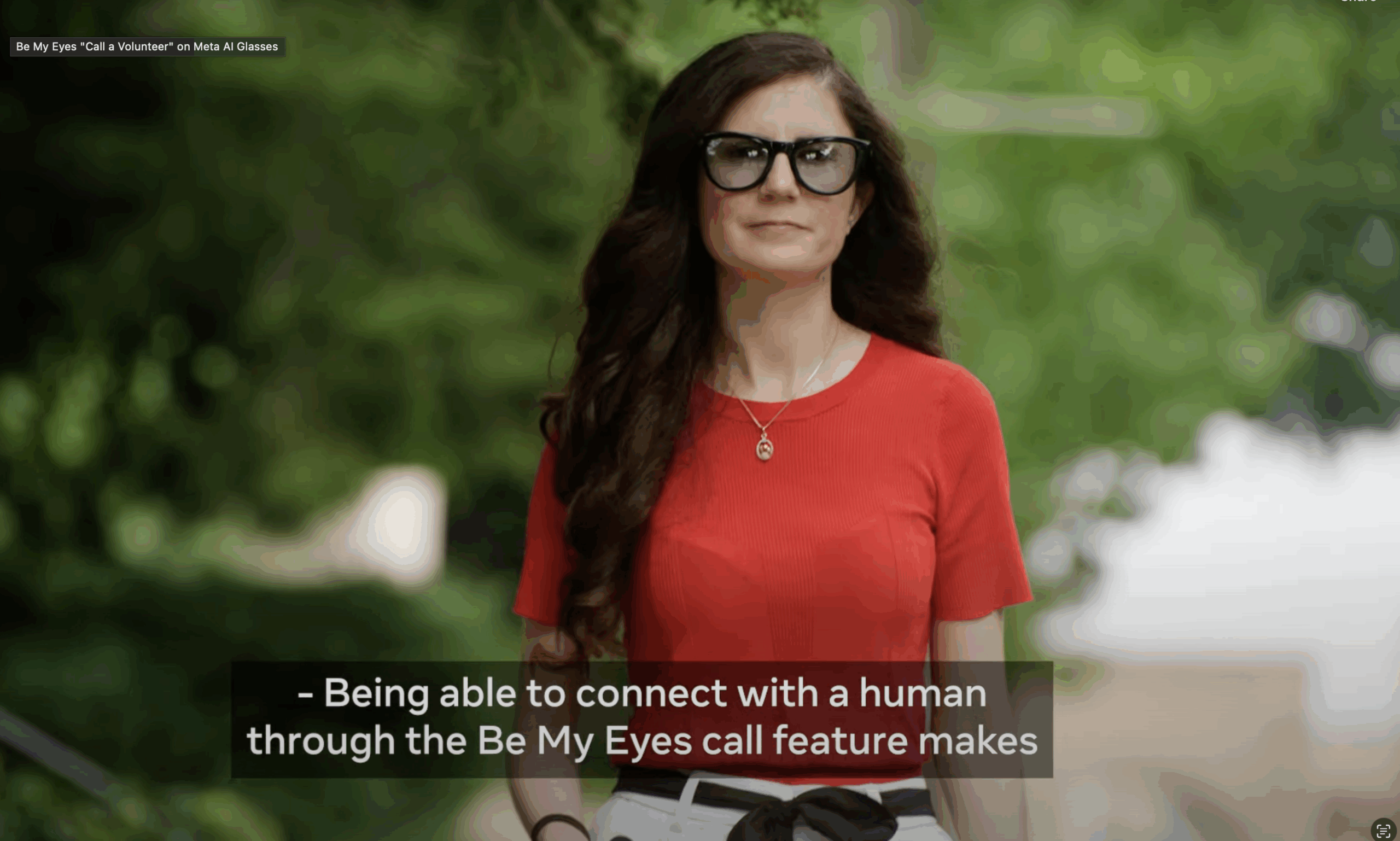 A woman with long, dark wavy hair and black-framed Meta AI glasses walks towards the camera. She is outdoors and is wearing a red, short-sleeved top and a gold pendant necklace, with blurred green foliage in the background.
