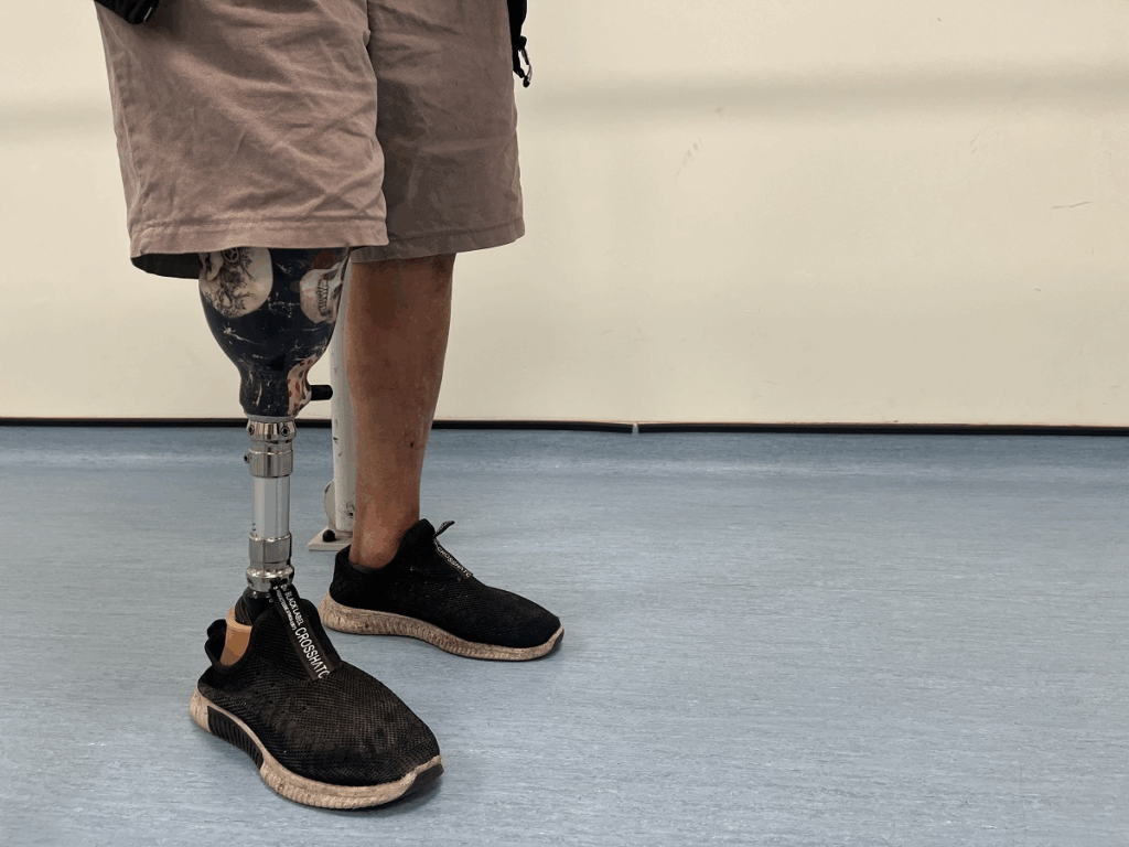 A close-up, low-angle shot of a person standing on a blue floor, showing their right leg which is a prosthetic limb from below the knee. The socket of the prosthetic is decorated black and white