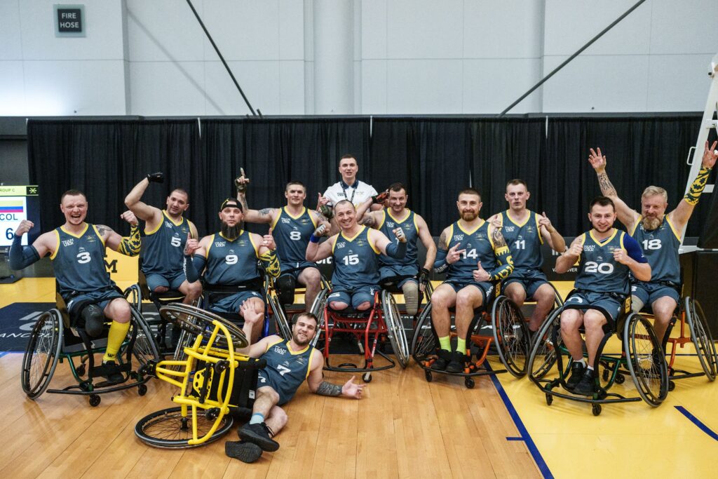 A team of male wheelchair basketball players in matching blue and yellow uniforms poses for a celebratory group photo on a basketball court. Many are smiling, flexing their biceps, and raising their arms in triumph.