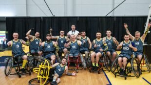 A team of male wheelchair basketball players in matching blue and yellow uniforms poses for a celebratory group photo on a basketball court. Many are smiling, flexing their biceps, and raising their arms in triumph.
