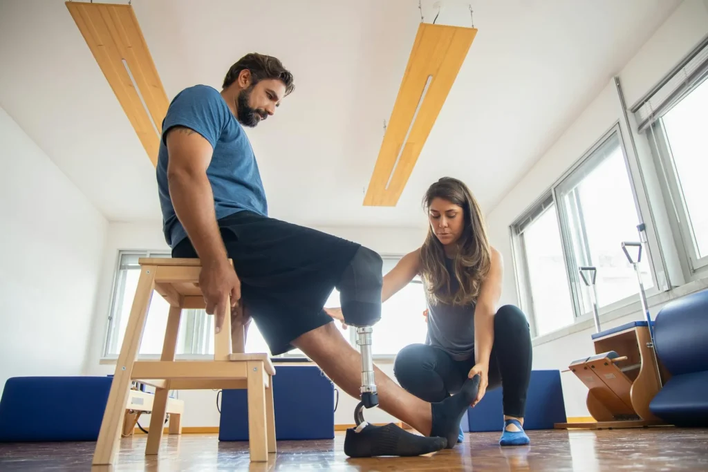A man with a prosthetic right leg sits on a wooden stool in a clinic while a female therapist crouches down to examine and adjust his prosthetic foot