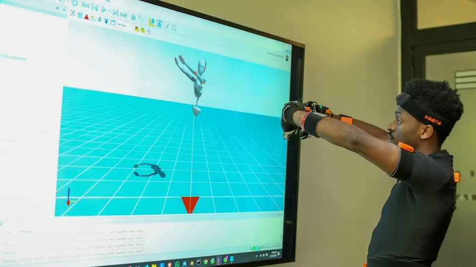Elly Savatia wearing motion capture gloves with orange sensors demonstrates sign language while facing a large monitor displaying a 3D avatar mimicking the hand movements in a virtual environment with a turquoise grid background