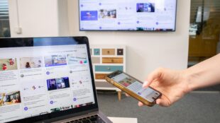 A hand with a polished fingernail holds a gold-cased smartphone directly in front of an open laptop. The phone screen is active and shows a webpage with an image and text. Directly behind the phone, the laptop screen displays a grid of content cards. In the background, a multi-colored dresser is visible,Above the dresser, a large wall-mounted flat-screen TV is present, also displaying the same complex grid content from a distance, mirroring the other two devices. The lighting is diffused, and the scene emphasizes seamless multi-screen content viewing. A window is in the background. The laptop keyboard is in the immediate foreground.