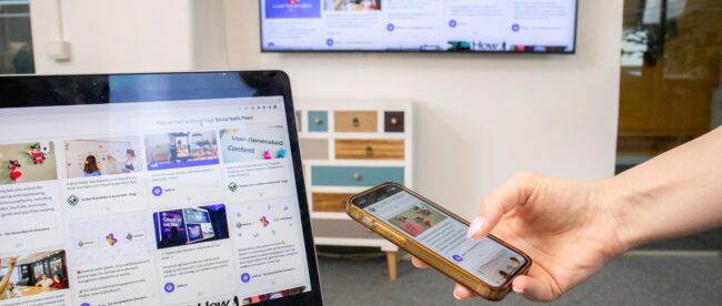 A hand with a polished fingernail holds a gold-cased smartphone directly in front of an open laptop. The phone screen is active and shows a webpage with an image and text. Directly behind the phone, the laptop screen displays a grid of content cards. In the background, a multi-colored dresser is visible,Above the dresser, a large wall-mounted flat-screen TV is present, also displaying the same complex grid content from a distance, mirroring the other two devices. The lighting is diffused, and the scene emphasizes seamless multi-screen content viewing. A window is in the background. The laptop keyboard is in the immediate foreground.