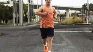 A man with Down syndrome, athlete Chris Nikic, running confidently down a paved road in a bright orange tank top and the orange adidas Supernova Rise 3 Adaptive running shoes.
